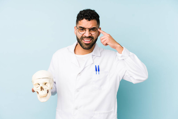 Young doctor latin man holding a skull isolated showing a disappointment gesture with forefinger. - Photo, Image