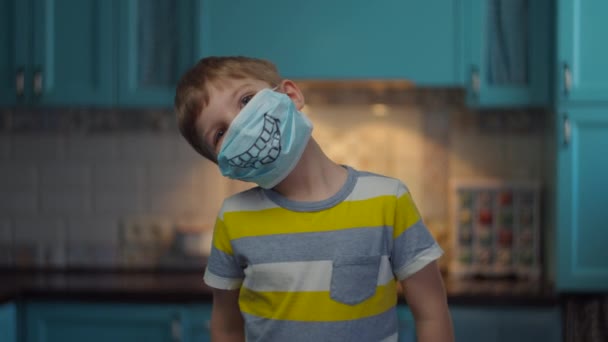 Portrait of boy in medical mask with smile looking at camera. Kid in funny medical mask at home in self isolation from virus. Stop epidemic. - Video, Çekim