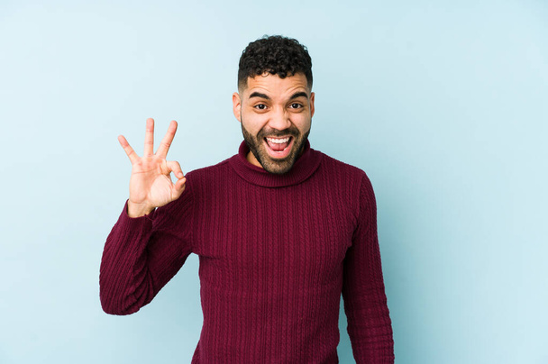 Young mixed race arabic man isolated winks an eye and holds an okay gesture with hand. - Photo, Image