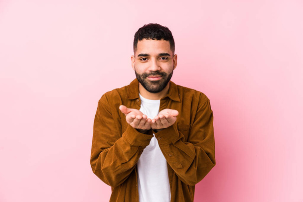 Young latin man against a pink background isolated holding something with palms, offering to camera. - Fotoğraf, Görsel