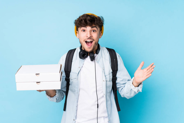 Jeune étudiant arabe tenant des pizzas isolées recevant une agréable surprise, excitée et levant la main
. - Photo, image