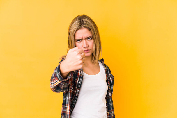 Giovane donna caucasica bionda isolata su uno sfondo giallo mostrando pugno alla macchina fotografica, espressione facciale aggressiva
. - Foto, immagini