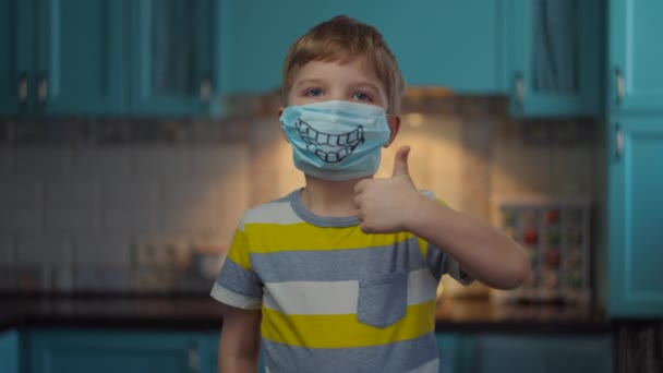 Portrait of boy in medical mask with smile looking at camera showing thumbs up. Kid in funny medical mask at home in self isolation from virus. Stop epidemic. - 映像、動画