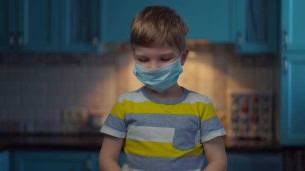 Blonde boy in medical mask on face measures temperature with infrared thermometer looking at camera. Kid in medical mask at home in self isolation from virus. Stop epidemic. - Záběry, video
