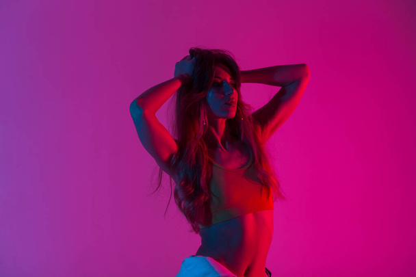 Πανέμορφο σύγχρονο μοντέλο μοντέρνα κομψή γυναίκα σε ένα σέξι μπλουζάκι με ένα τέλειο σώμα ισιώνει μακριά μαλλιά και απολαμβάνει ένα φωτεινό νέον κόκκινο φως. Ελκυστικό κορίτσι που ποζάρει σε ένα δωμάτιο με πολύχρωμο ντίσκο χρώμα. - Φωτογραφία, εικόνα