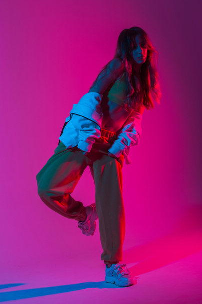 Trendy young woman dancer in a youth fashion tracksuit in stylish sneakers dancing hip-hop in the studio with bright pink neon light. Modern girl enjoys a dance in the studio with multi-colored color. - Fotografie, Obrázek