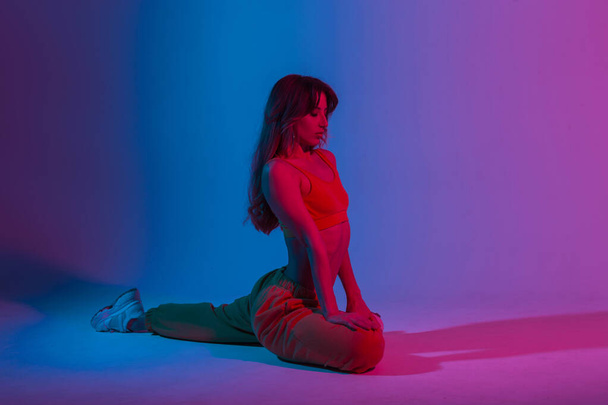 Nowoczesna wysportowana młoda piękna kobieta w odzieży młodzieżowej robi fitness w pokoju z niesamowitym jasnym niebieskim światłem. Sport dziewczyna robi ćwiczenia jogi w studio z neon multi-kolorowy kolor. - Zdjęcie, obraz