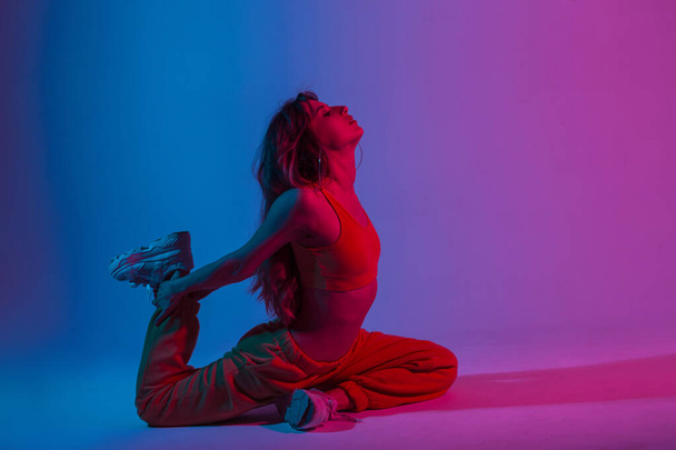 Professional cool young woman in fashionable sportswear doing fitness in a room with multicolored neon color. Attractive girl yoga instructor training in the studio with bright pink disco light. - 写真・画像