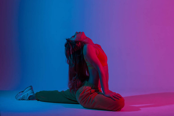 Młoda kobieta instruktor jogi w modnej odzieży sportowej robi ćwiczenia rozciągające z wielokolorowym neonowym tle koloru. Profesjonalna dziewczyna robi fitness w studio z jasnoniebieskim świetle disco. - Zdjęcie, obraz