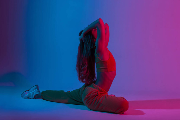 Mooie jonge vrouw in een stijlvolle outfit die fitness doet in de studio met een helder neon licht. Atletisch aantrekkelijk meisje mode model doen yoga oefeningen binnen met multi-gekleurde glitter. - Foto, afbeelding