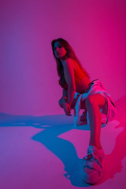 Sporty pretty young woman dancer with stylish hoodie in a top in sneakers sits in a room with bright blue-violet light.Sexy girl model posing in trendy wear indoors with neon pink color in disco style - 写真・画像
