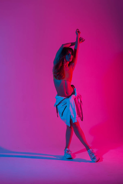 Mujer joven sexy delgada en ropa juvenil de moda posando en una habitación con color rosa y azul en estilo disco. Hermosa chica con estilo está de pie en el estudio con una luz de varios colores con estilo increíble
. - Foto, Imagen
