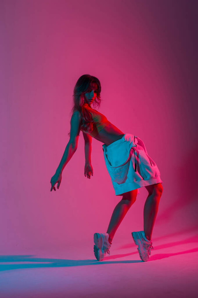 Sports young woman dancer in a youth fashion toh in hoodie in stylish sneakers dancing hip-hop in the studio with bright pink neon light.Cool girl enjoys a dance with multi-colored color in the studio - Fotó, kép