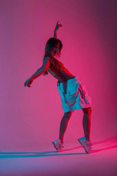 Μοντέρνο μοντέλο νεαρή γυναίκα στα ρούχα μόδας σε παπούτσια που ποζάρουν στο στούντιο με πολύχρωμο υπεριώδες χρώμα. Σύγχρονη χορεύτρια κορίτσι χορεύει σε ένα δωμάτιο με φωτεινά ροζ νέον φώτα σε στυλ ντίσκο. - Φωτογραφία, εικόνα
