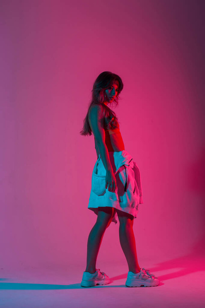 Атлетичная молодая женщина в винтажном топе в модной толстовке позирует в комнате с разноцветным цветом в стиле диско. Крутая девушка в модной одежде стоит в комнате с ярким неоновым сине-фиолетовым светом
 - Фото, изображение