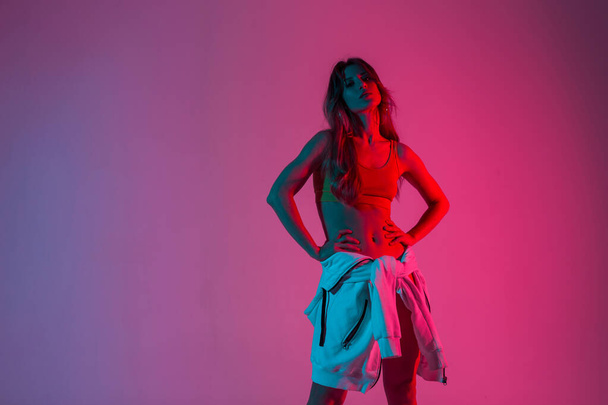 Αθλητική νεαρή γυναίκα σε ένα vintage top σε hoodie θέτει σε ένα δωμάτιο σε ένα ροζ πολύχρωμο φόντο. Σέξι κορίτσι μόδας μοντέλο σε μοντέρνα ρούχα στέκεται στο στούντιο με φωτεινό νέον ροζ φως. - Φωτογραφία, εικόνα