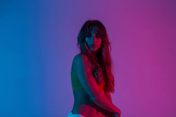 Atletische jonge vrouw met lang haar in een modieuze top poseert in een kamer op een blauwe kleurrijke achtergrond. Sexy meisje mode model in trendy slijtage staat in de studio met heldere neon roze licht. - Foto, afbeelding