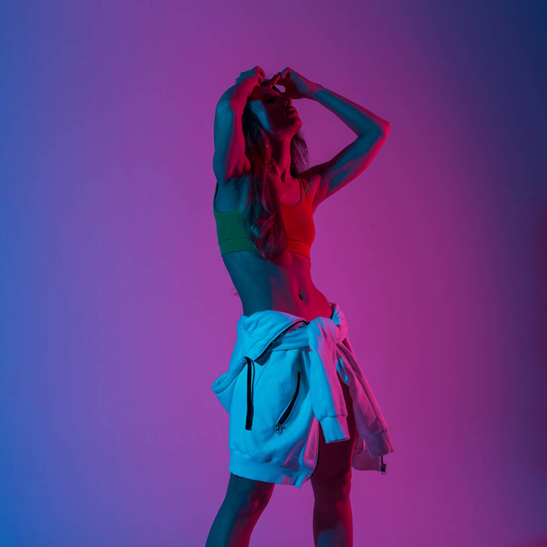 Tyylikäs nuori malli nainen muodikkaissa vaatteissa huppari seisoo huoneessa, jossa on kirkkaat neonvalot. Moderni trendikäs tyttö poseeraa studiossa vaaleanpunainen-sininen väri. Muotirouvat. Luova käsite
. - Valokuva, kuva
