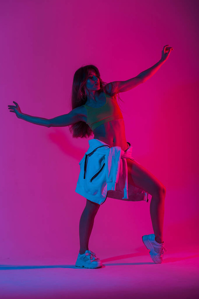 Glamorosa bailarina joven en ropa de moda deportiva juvenil posando en una habitación con estilo disco de color rosa multicolor. Atractiva modelo de moda chica está de pie en el estudio con una luz de neón brillante
 - Foto, Imagen