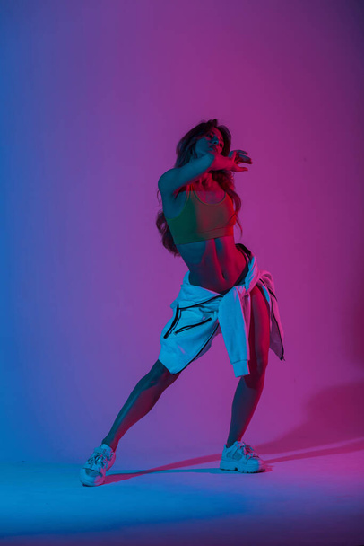 Young woman dancer in stylish youth clothes in gym shoes dancing and posing in a room with bright neon blue-pink color. Sports girl enjoys a dance in the studio with multi-colored ultraviolet light. - Photo, image