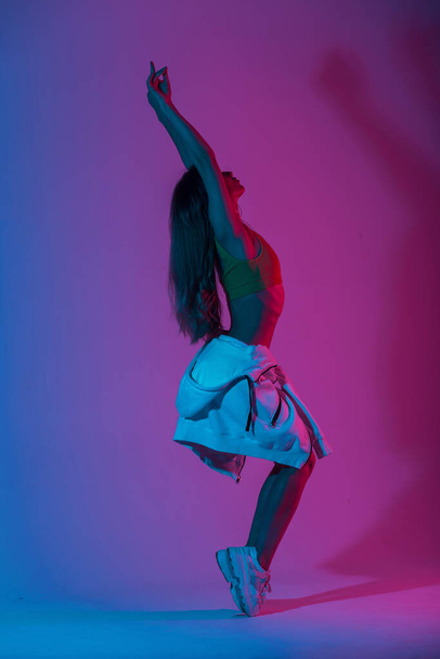 Trendy jonge vrouw danser in een jeugd mode dragen in stijlvolle sneakers dansen in de studio met fel roze neon licht. Sexy modern meisje geniet van een dans in de kamer met gekleurde kleur in disco stijl. - Foto, afbeelding