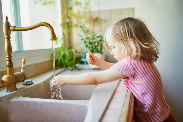 Adorable toddler girl washing her hands under kitchen sink. Little girl having fun with water. Hygiene and skin care for kids - Fotografie, Obrázek