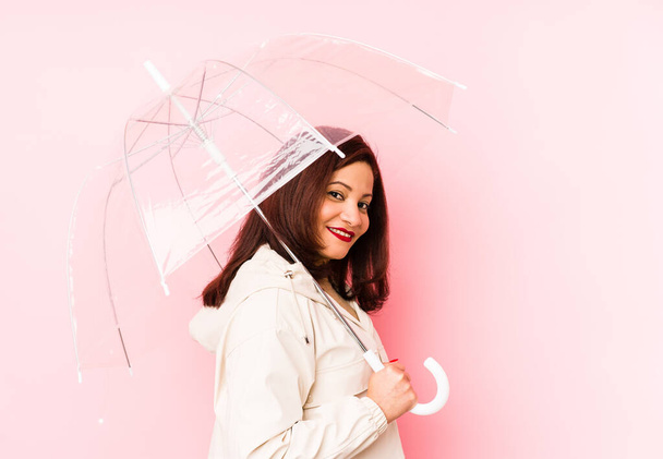 Middelbare leeftijd latijn vrouw draagt een paraplu geïsoleerde kijkt opzij glimlachend, vrolijk en aangenaam. - Foto, afbeelding