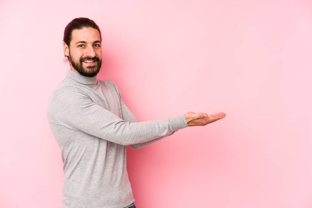 Young long hair man isolated on a pink background holding a copy space on a palm. - Foto, imagen