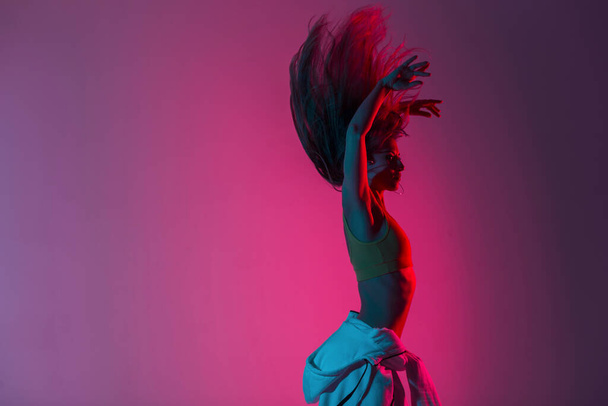 Modern young woman in fashionable clothes dancing in studio with pink color background. Beautiful trendy girl the dancer enjoys dancing in the room with colored light in style disco. Art dance concept - Valokuva, kuva