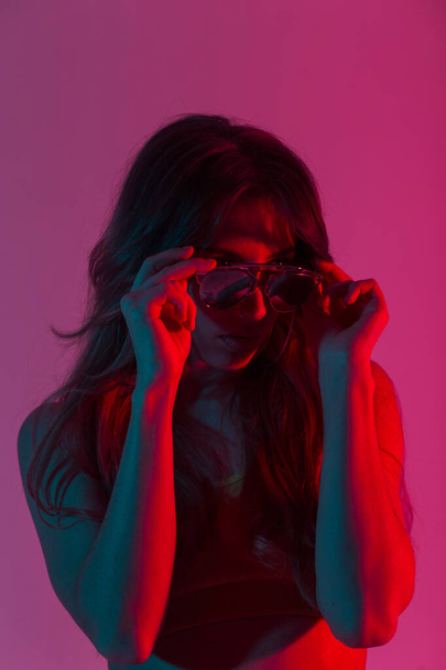 Stylowy model mody młodej kobiety z seksownymi ustami w koszuli modnej prostuje modne okulary przeciwsłoneczne w pomieszczeniach w kolorze różowo-czerwonym. Portret nowoczesna dziewczyna hipster w studio z kolorowymi światłami w stylu disco. - Zdjęcie, obraz