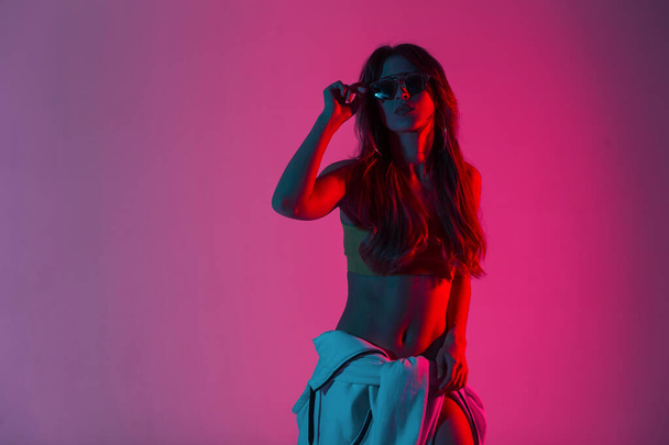Pretty slim young woman in a top with a sexy body with long hair puts on fashionable sunglasses in room with neon pink color background.Modern hipster girl posing indoors with bright red amazing light - Photo, Image