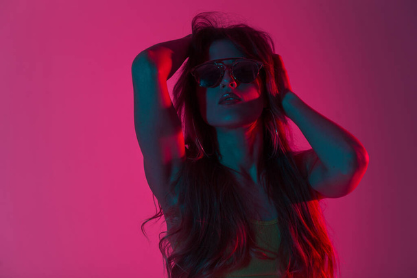 Fashionable young woman model in stylish top straightens long hair indoors with pink-red color background. Portrait girl hipster in trendy sunglasses with sexy lips in studio with colorful lights. - Foto, Bild