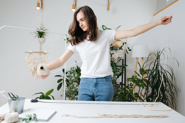 The girl unwinds the cotton rope, prepares to weave a macrame at home in a beautiful interior filled with flowers and plants - Фото, изображение