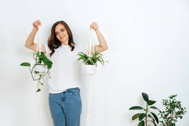 A pretty woman holds a pot of macrame with beautiful plants. She's wearing a white T-shirt and jeans, against a white wall - Valokuva, kuva