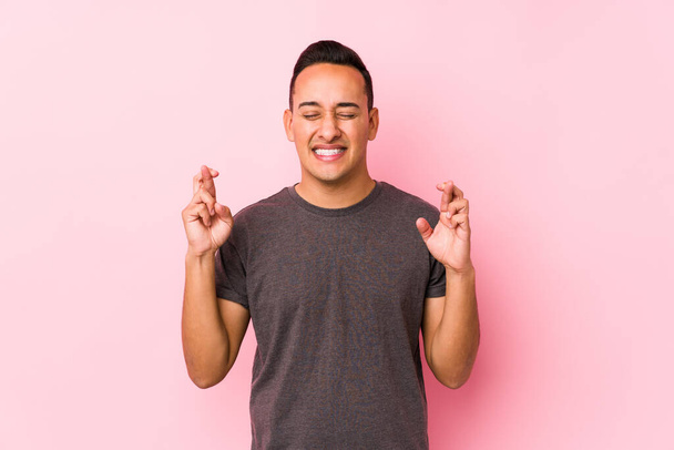Yooung latin man posando en un fondo rosadocruzando los dedos para tener suerte
 - Foto, Imagen