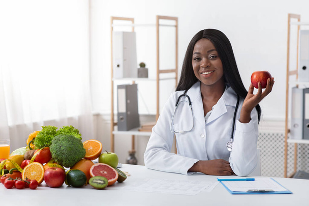 Afrykański amerykański uśmiechnięty lekarz dietetyk siedzi przy biurku, trzymając jabłko - Zdjęcie, obraz