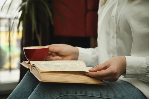 Woman with cup of coffee reading book indoors, closeup - Photo, Image