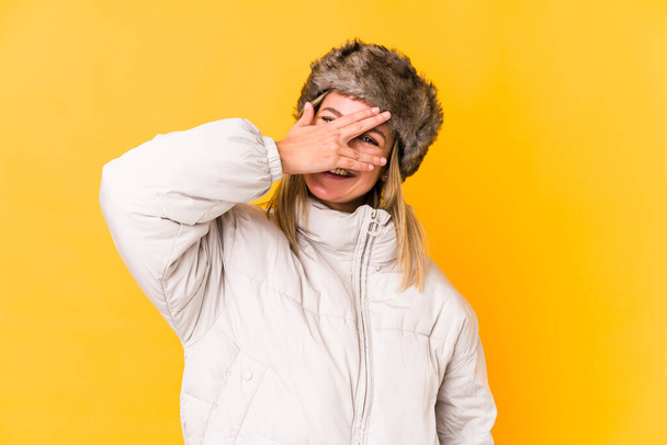 Young blonde caucasian woman wearing a winter clothes isolated blink at the camera through fingers, embarrassed covering face. - Photo, Image