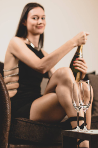 Classy young woman wearing a fancy dress, sitting on couch in a lounge, opening a champagne bottle. Royalty free stock photo. - Photo, Image