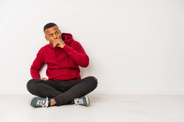 Young latin man sitting on the floor isolated thoughtful looking to a copy space covering mouth with hand. - Photo, Image