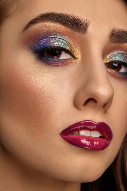 Brunette female with luxury makeup and perfect skin is looking at you. Multi-colored eyeshadow, false eyelashes, glossy burgundy lips. Professional maquillage. Close up - Foto, Bild