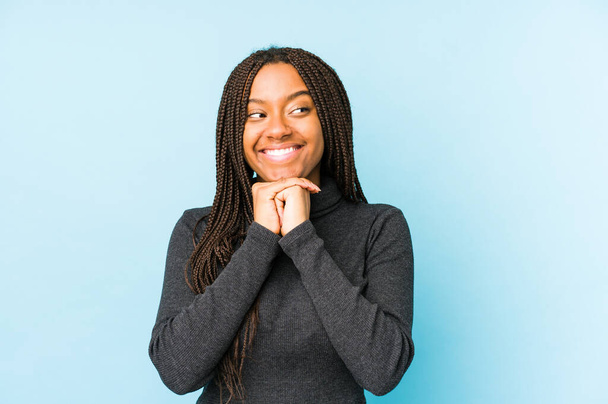 Junge afrikanisch-amerikanische Frau auf blauem Hintergrund hält die Hände unter dem Kinn, blickt glücklich zur Seite. - Foto, Bild