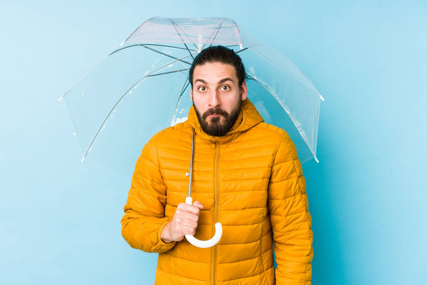 Νεαρός άνδρας με μακριά μαλλιά κοιτάζει κρατώντας μια ομπρέλα απομονωμένη ανασηκώνει ώμους και ανοιχτά μάτια συγχέεται. - Φωτογραφία, εικόνα