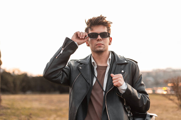 Fashionable young hipster man in vintage leather oversized black jacket with backpack straightens sunglasses outdoors. American guy in trendy casual clothes posing in nature. Retro fashion. - Photo, Image
