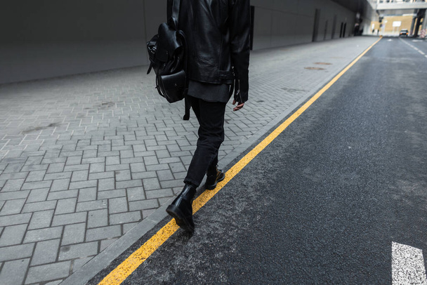 Современный молодой человек в стильной куртке в винтажных джинсах в кожаных модных сапогах с модным черным рюкзаком выходит на улицу. Парень в чёрной юности путешествует по городу. Желтая линия
 - Фото, изображение
