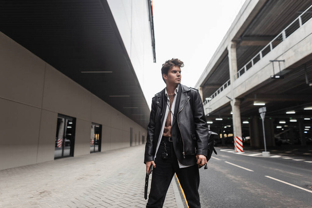 Bello europeo giovane hipster uomo con acconciatura in giacca di pelle alla moda in jeans alla moda con zaino alla moda passeggiate per strada vicino alla strada. attraente ragazzo in nero giovani vestiti all'aperto
 - Foto, immagini