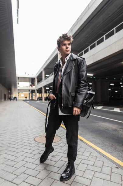 Modern young hipster man in trendy black casual wear poses in the city near the road. Stylish modern urban guy model in a stylish leather jacket oversize in trousers in boots outdoors. Street fashion. - Foto, immagini