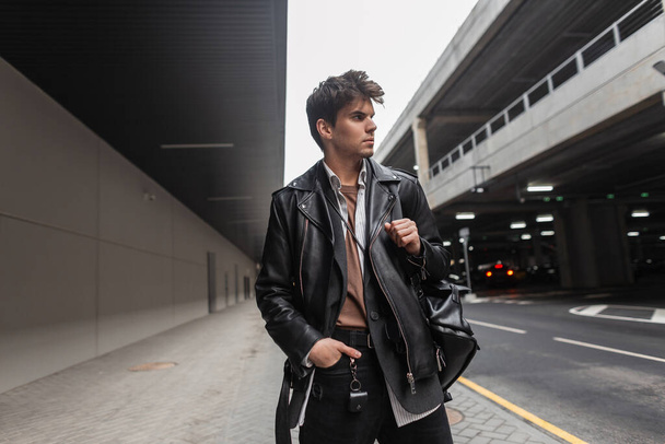 Модный молодой человек в модной кожаной черной куртке в рубашке в штанах со стильным рюкзаком с прической, позирующей возле дороги в городе. Европейская модель парня в молодежной повседневной одежде
. - Фото, изображение