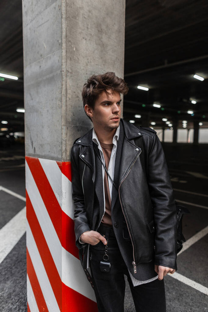 Ernstig trendy jongeman model in een jeugd oversized modieuze zwarte leren jas met een stijlvolle kapsel staat in de buurt van een grijze kolom op een parkeerplaats in de stad. Knappe hipster moderne man in de straat - Foto, afbeelding