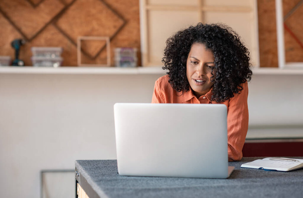 Jonge vrouw met krullend haar zitten op een werkbank in haar foto inlijsten studio werken online met een laptop - Foto, afbeelding